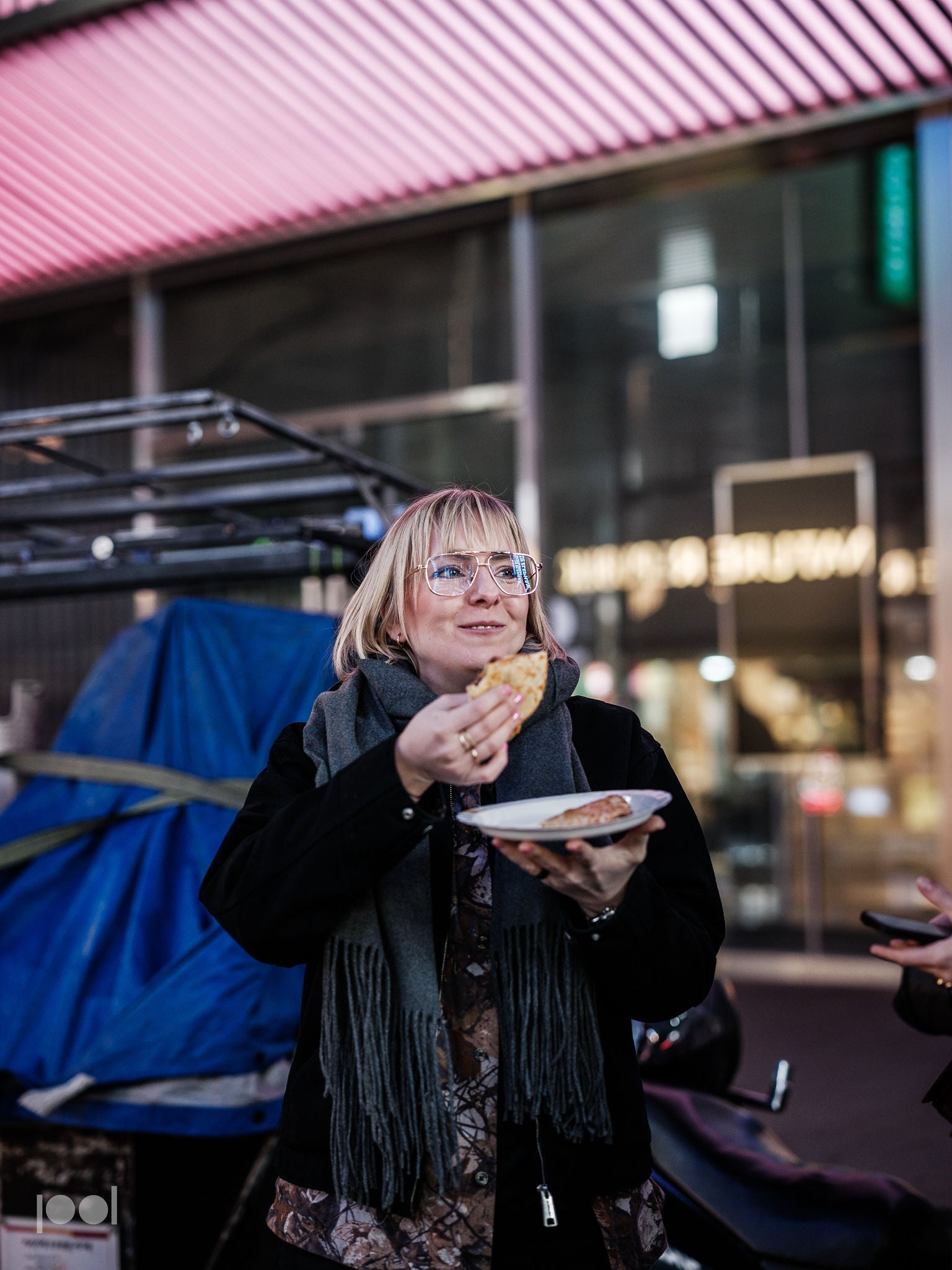 A woman with glasses and a scarf smiles while enjoying a meal served on a plate, outdoors in an urban environment illuminated by neon lights and blurred shop signs. The scene reflects the joy of dining with precision-engineered cookware, inspired by Fissler’s tradition of quality, durability, and timeless German design.