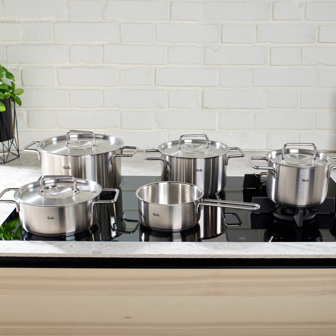 Fünf Fissler Edelstahltöpfe und -pfannen mit Deckeln stehen auf einer eleganten schwarzen Herdplatte und zeugen von deutscher Präzision und zeitlosem Design.